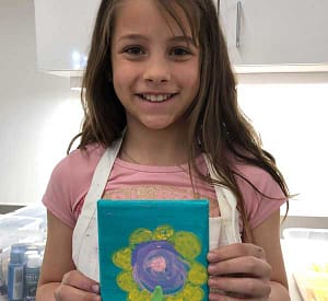 A smiling young female student artist showing her yellow petaled purple centered flower on a blue background painting.